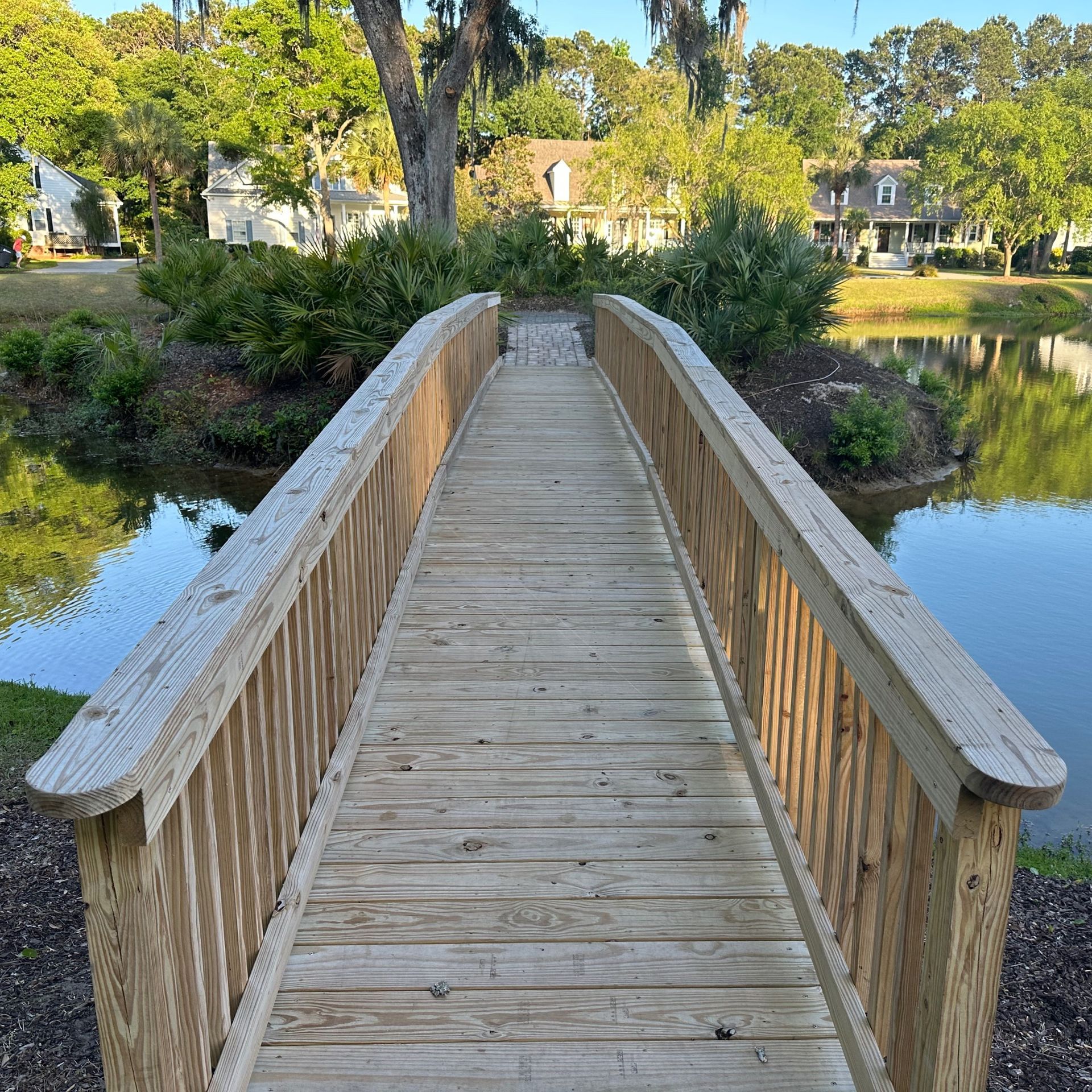 Wooden footbridge over a pond, leading to trees and houses in a sunny park.