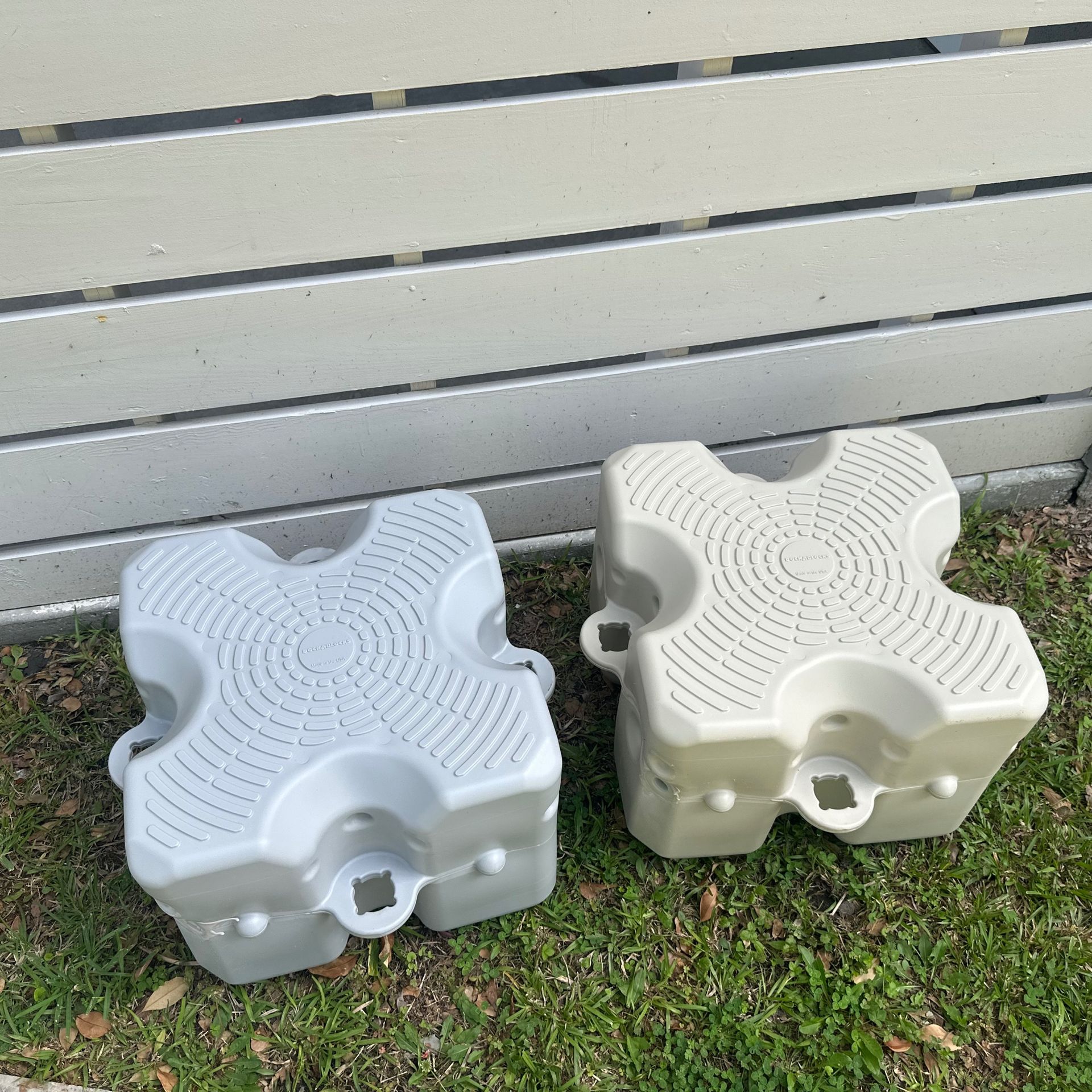 Two plastic step stools, one light blue and one off-white, sitting on grass beside a fence.