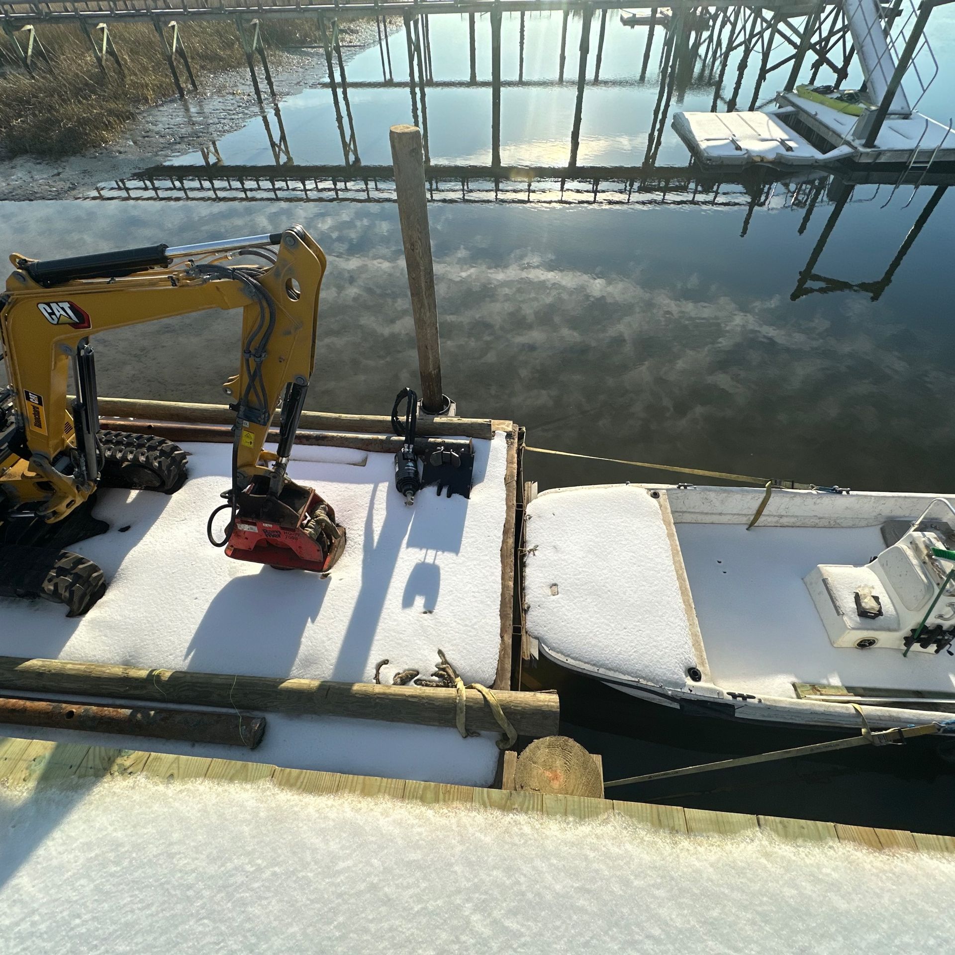 Snowy dock with a yellow excavator and a small boat beside frozen water.