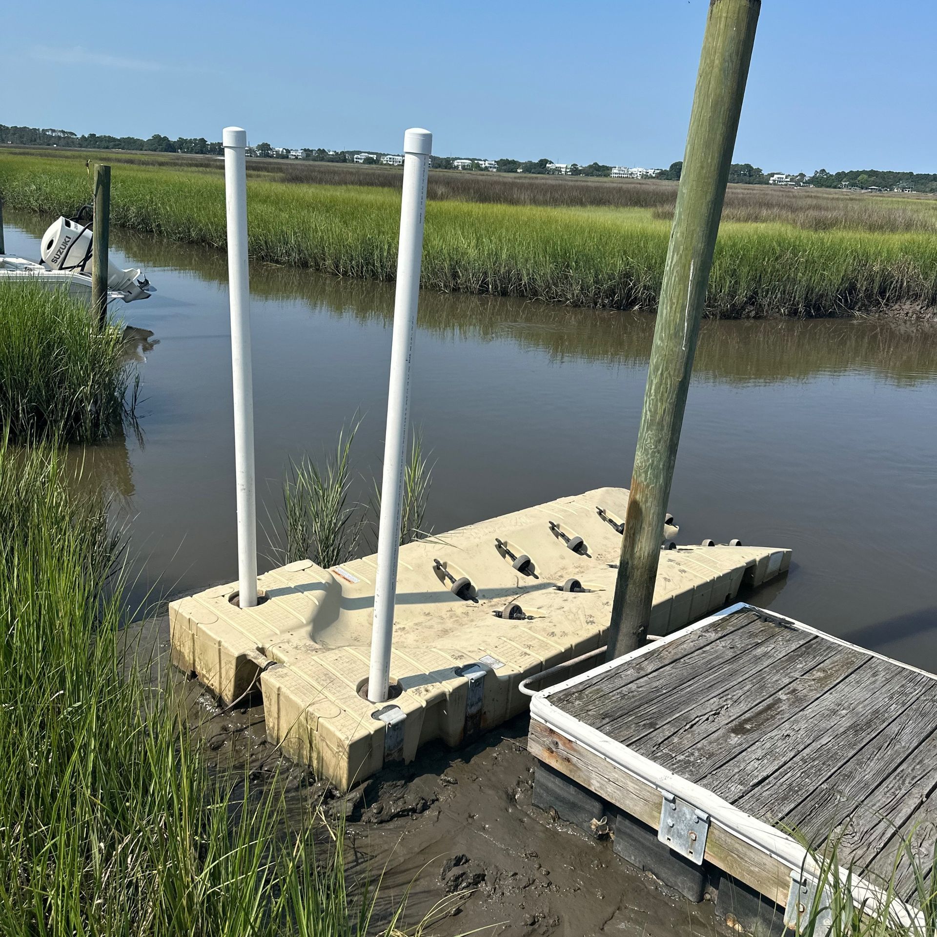 Small dock with posts by calm marsh water, beside reeds and a wooden platform under a clear sky