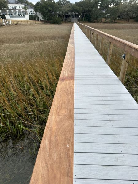 Wooden boardwalk stretching through marsh grass toward houses and trees under a cloudy sky
