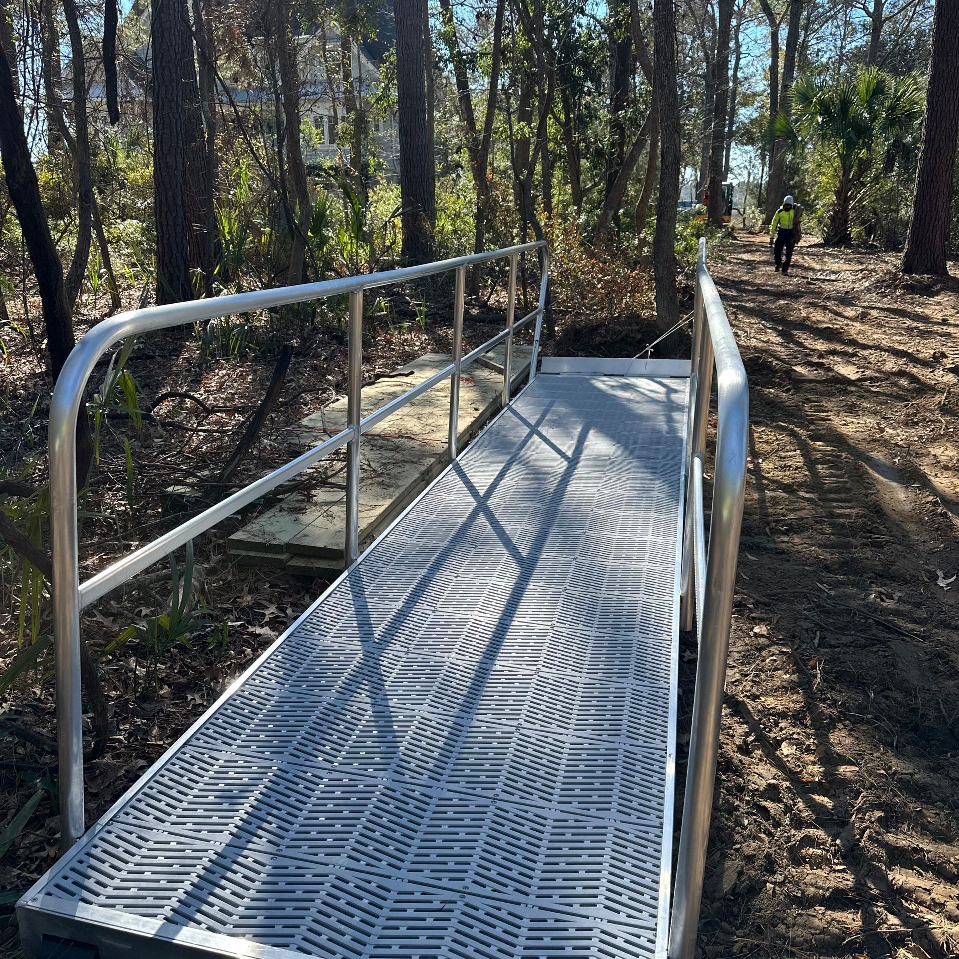 Metal accessibility ramp through a wooded area, with a person walking in the background.