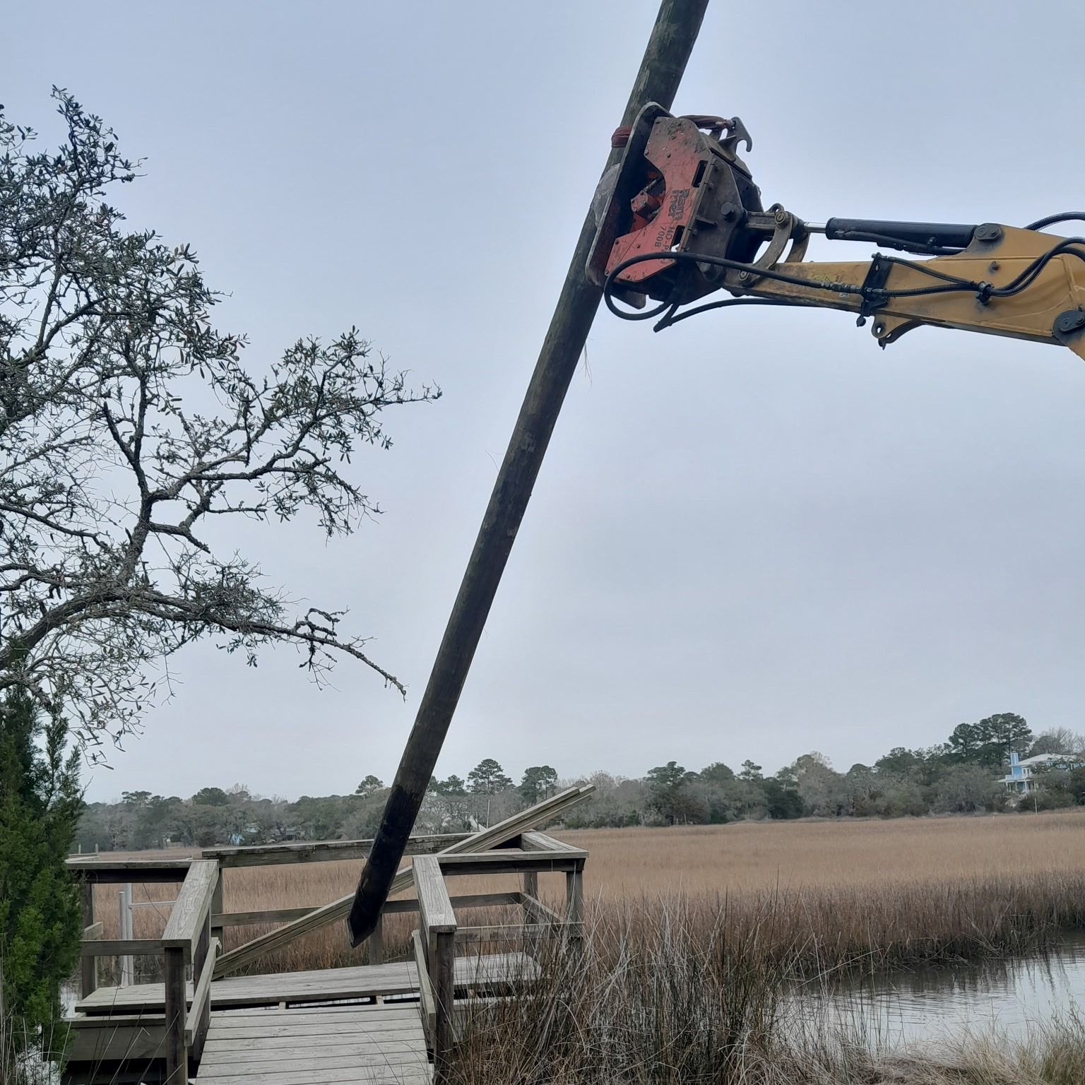 Excavator lifting a long black pipe over a marsh beside a wooden dock and trees
