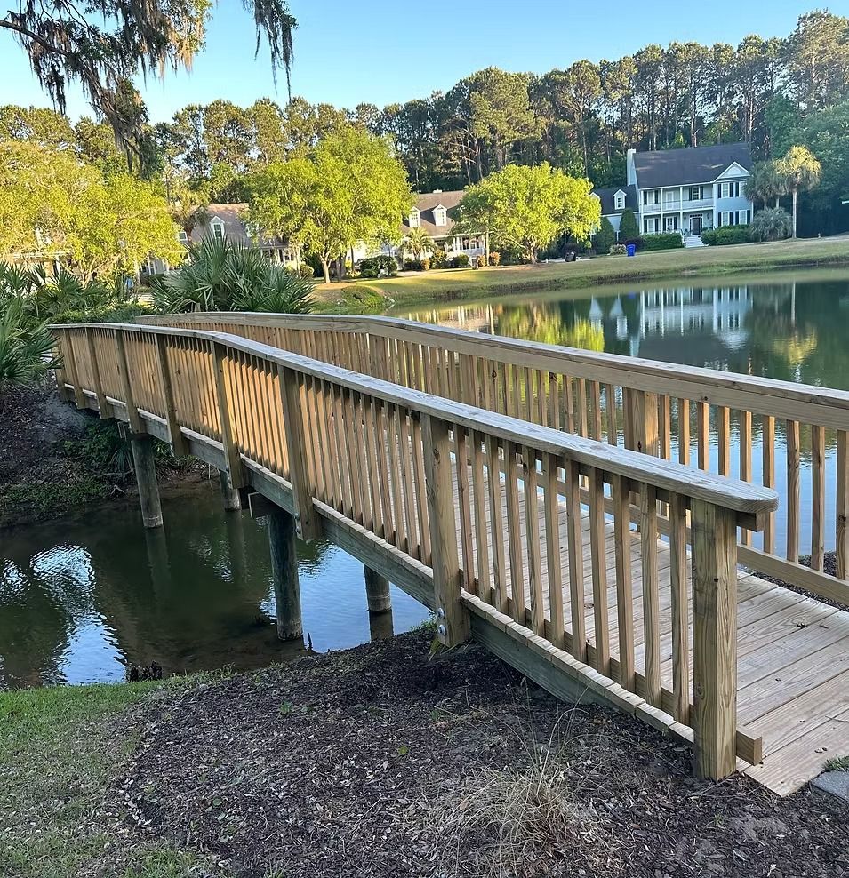 Wooden footbridge over a pond in a sunny park, with houses and trees in the background