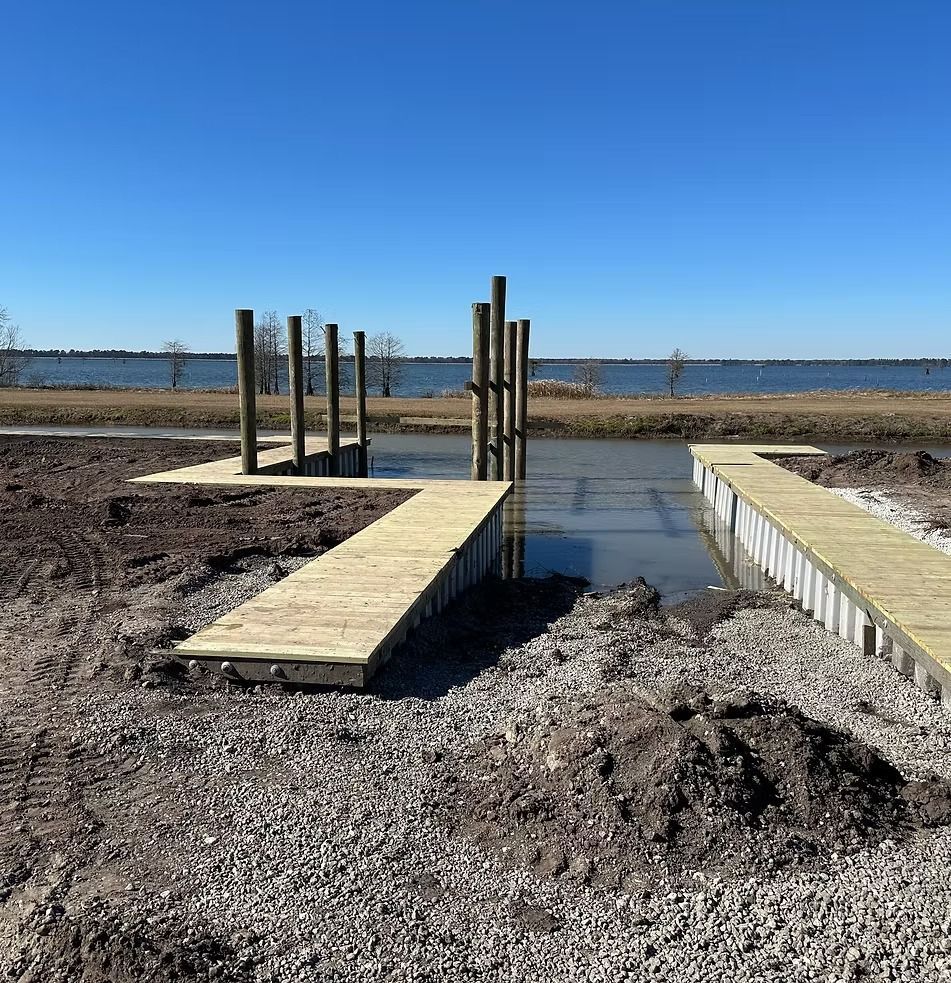Wooden dock under construction by a muddy shoreline, with a blue lake and clear sky in the background