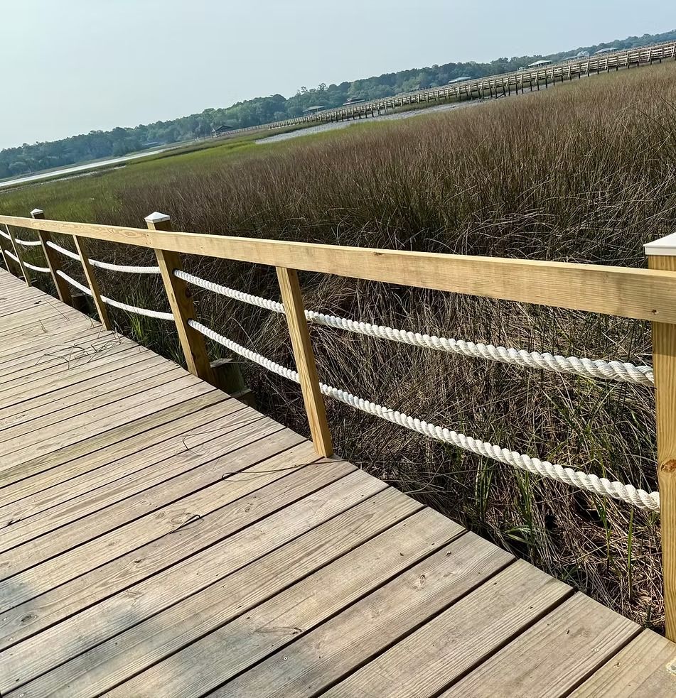 Wooden boardwalk beside tall marsh grass under a clear sky, with water and distant shoreline in view