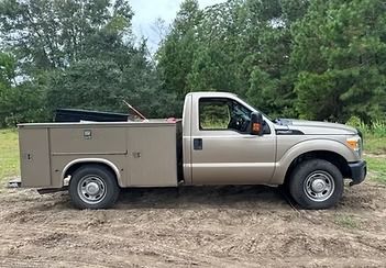 Tan pickup truck with a beige utility box parked on dirt near trees