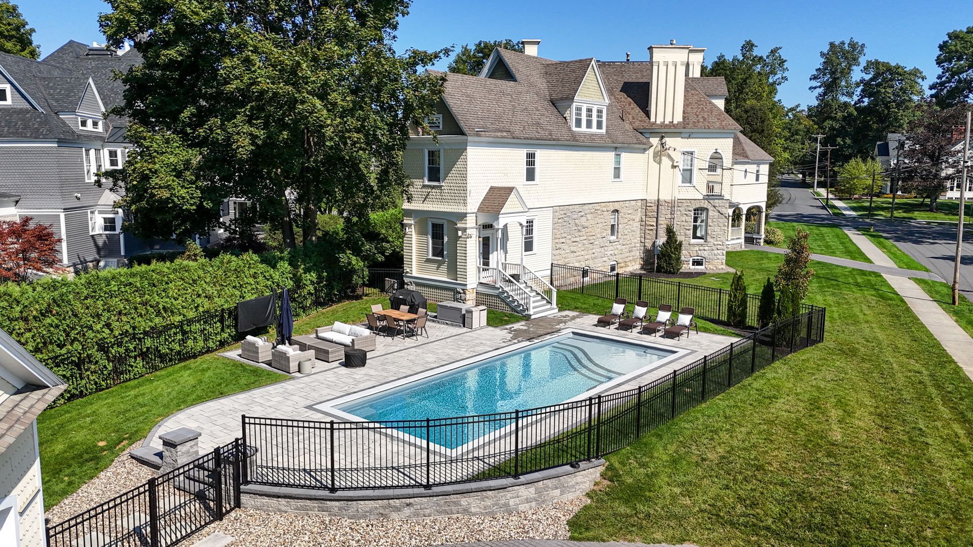 Stone house with a pool in a yard. Black fence surrounds pool with lounge chairs. Trees, green grass.