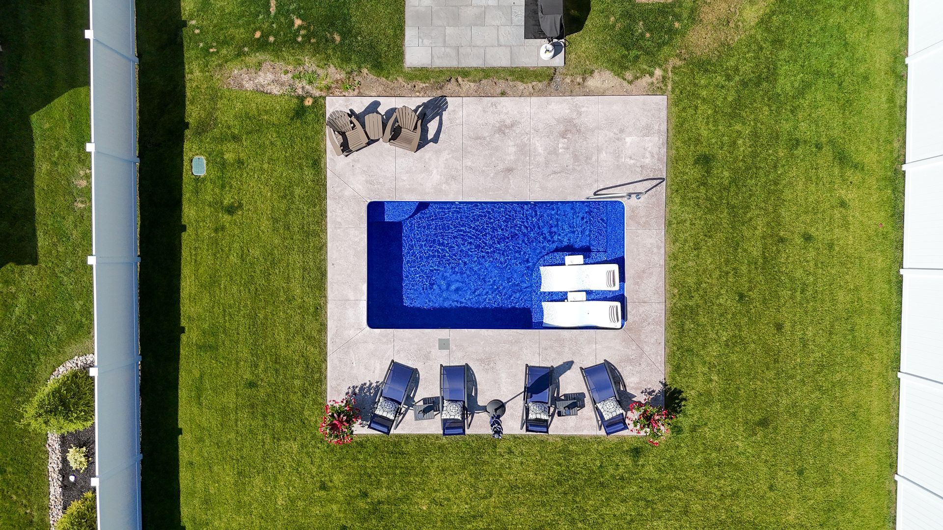 Overhead view of a rectangular blue pool surrounded by a concrete patio, lawn, and outdoor furniture.
