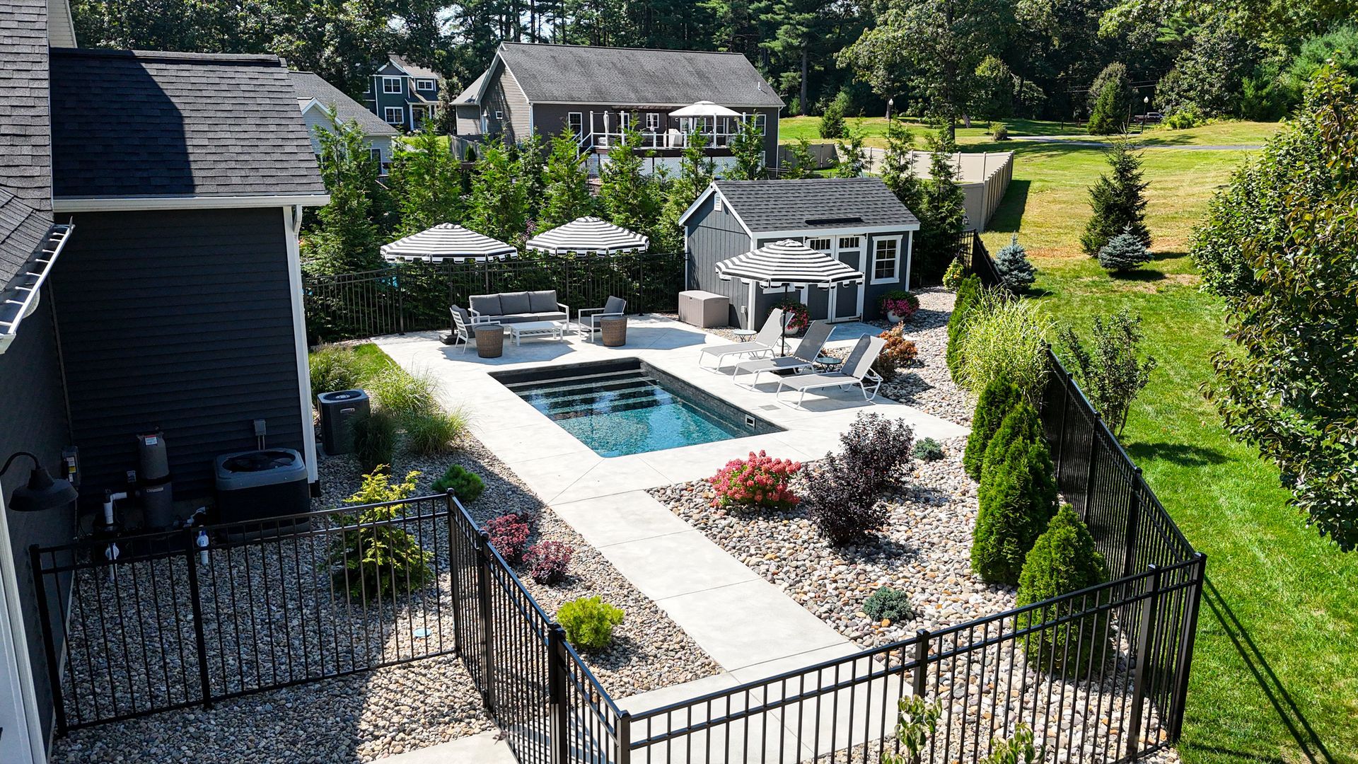 Backyard with a pool, patio, and small shed, with surrounding landscaping and houses in the distance.