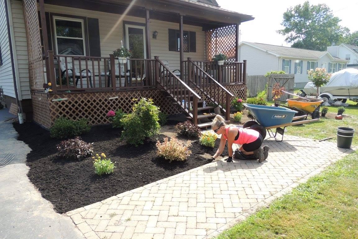 Person planting flowers beside a brick walkway in front of a house with a porch