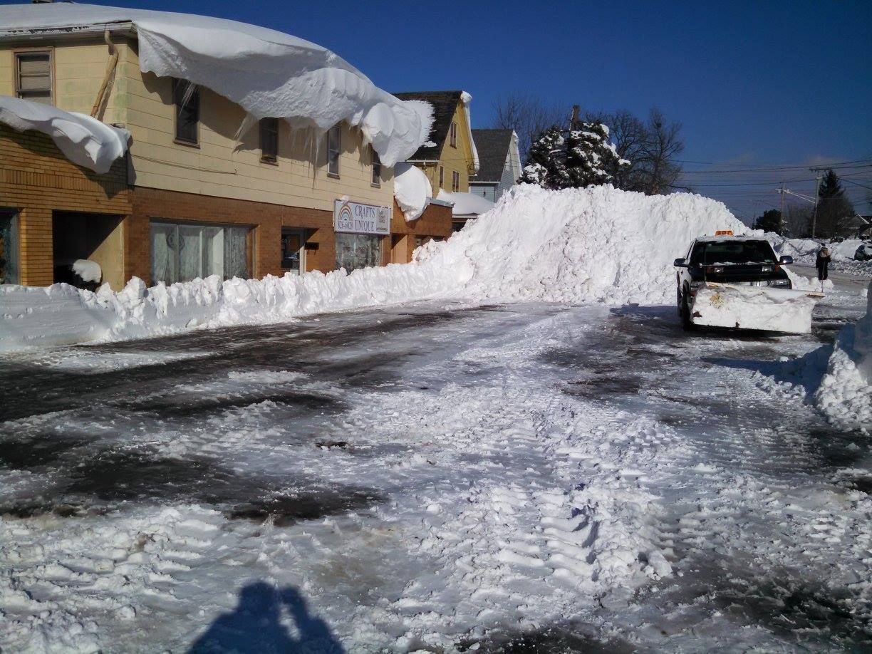 Snow-covered residential street with parked car, house, and large plowed snowbank under a clear blue sky