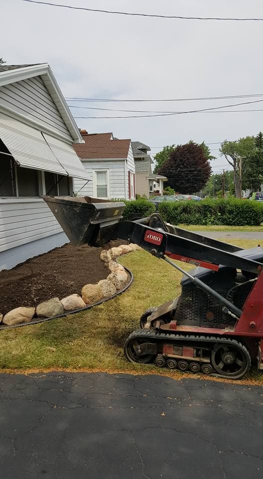 Mini excavator digging beside a house in a suburban yard, with rocks edging a small garden bed.