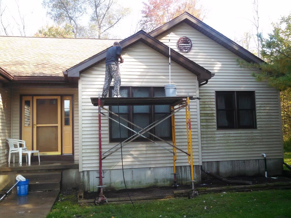 Worker on scaffold repairing house siding under front gable, with ladder and tools outside a gray home.