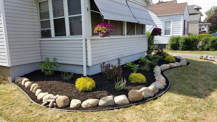 House corner with curved stone border, black mulch, yellow flowers, and hanging pink blooms by the porch