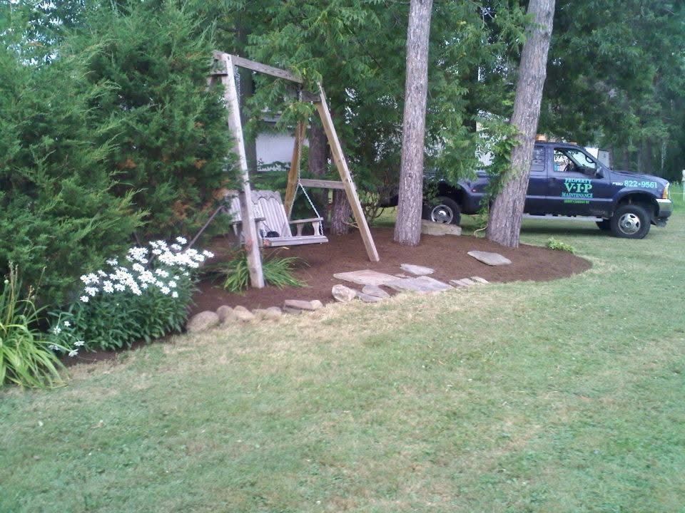 Wooden swing bench by trees in a grassy yard, with a green truck parked nearby.