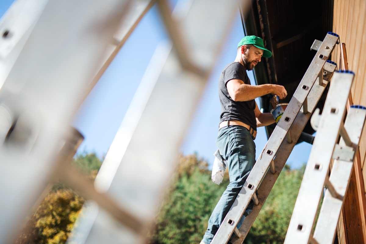 Man standing on a ladder leaning on the side of a house, applying paint from a can to the house