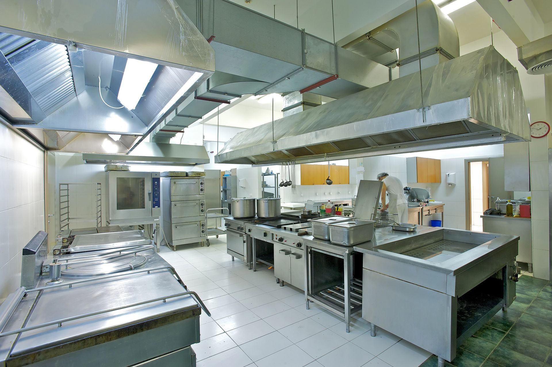 Cucina professionale con ripiani in acciaio inox, cappe aspiranti e pavimenti piastrellati in un ambiente di lavoro luminoso e pulito.