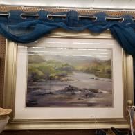 Framed landscape painting with a river, displayed under a blue draped curtain on a wall.