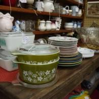 Green floral casserole dish on a shelf beside stacked colorful plates and white teapots in a kitchenware shop