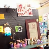 Craft booth with handmade items on a table, black display wall, and a sign reading “The Gen Designs”