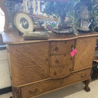 Wooden dresser with ornate carved details, topped with framed photos and decorative items, in a room.