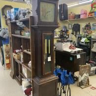 Antique grandfather clock in a cluttered thrift store aisle with shelves of assorted items