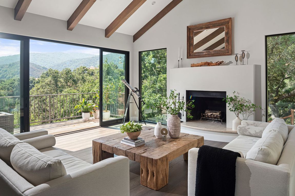 A bright sea ranch  living room with white sofas, a wooden coffee table, and a fireplace, featuring large windows overlooking trees.