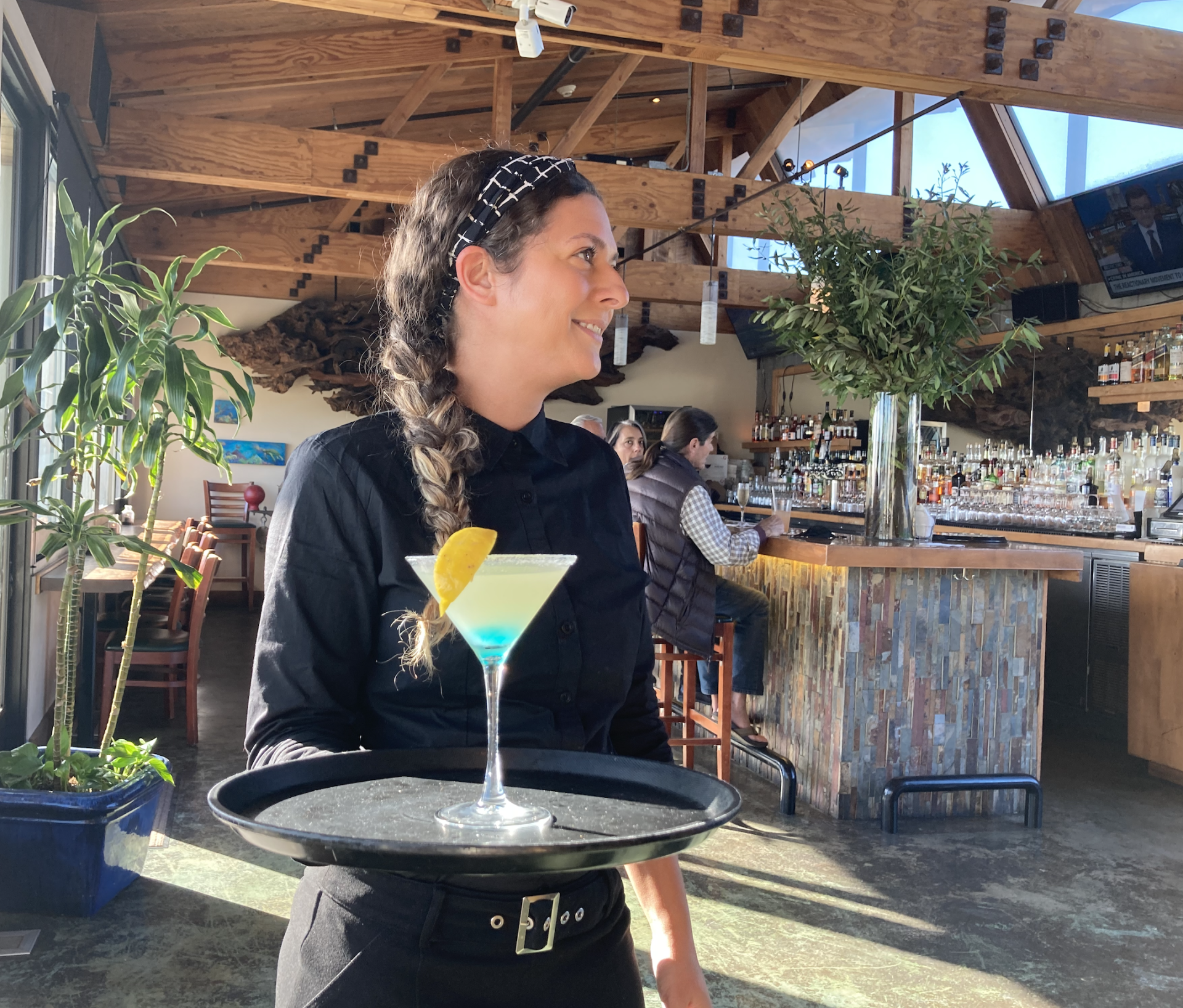 A server in a black uniform carries a martini glass with a lemon garnish on a tray in a sunlit restaurant.