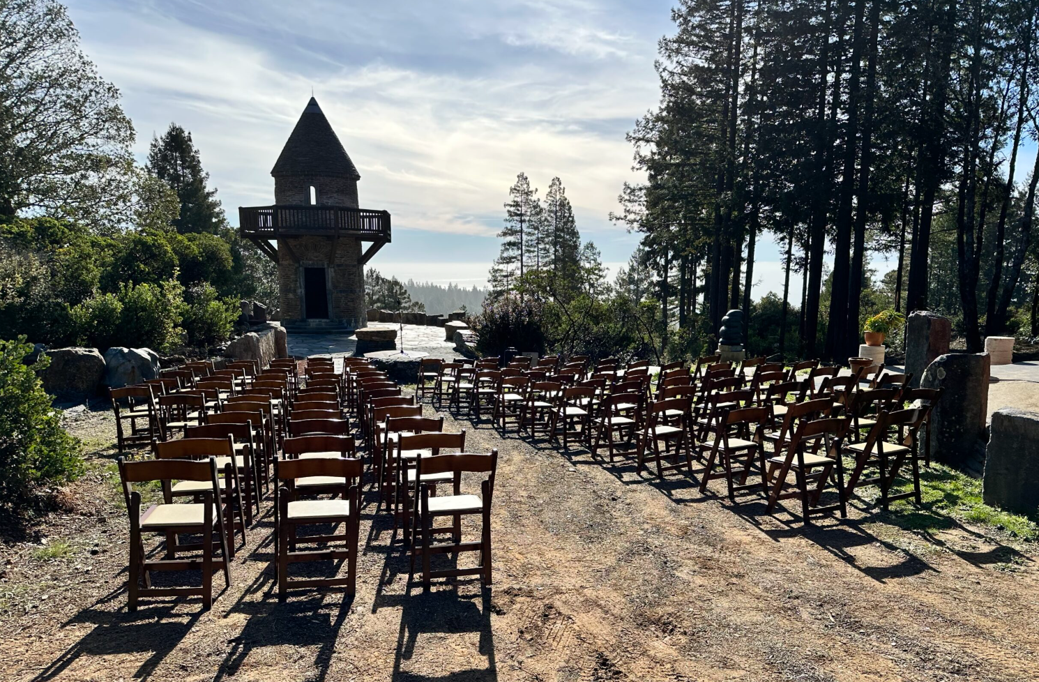 Mendocino Stone Ranch. Outdoor wedding ceremony setup with rows of wooden chairs facing a stone tower on a hilltop, surrounded by trees.