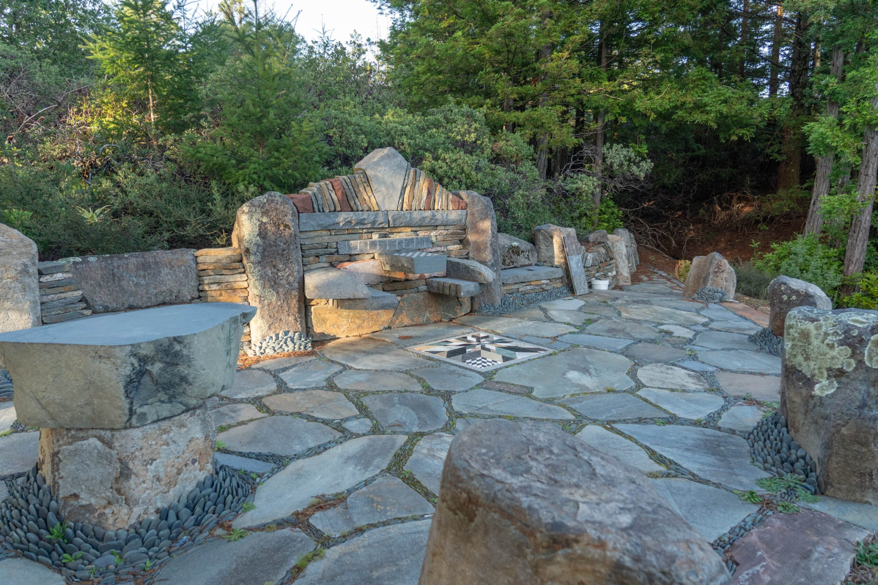 Mendocino Stone Ranch. A stone patio with a built-in stone bench, small fire pit, and woodland backdrop.