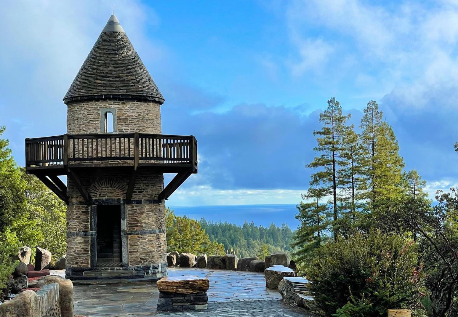 A stone lookout tower with a conical roof and wooden balcony, surrounded by trees and a stone patio under a cloudy sky.
