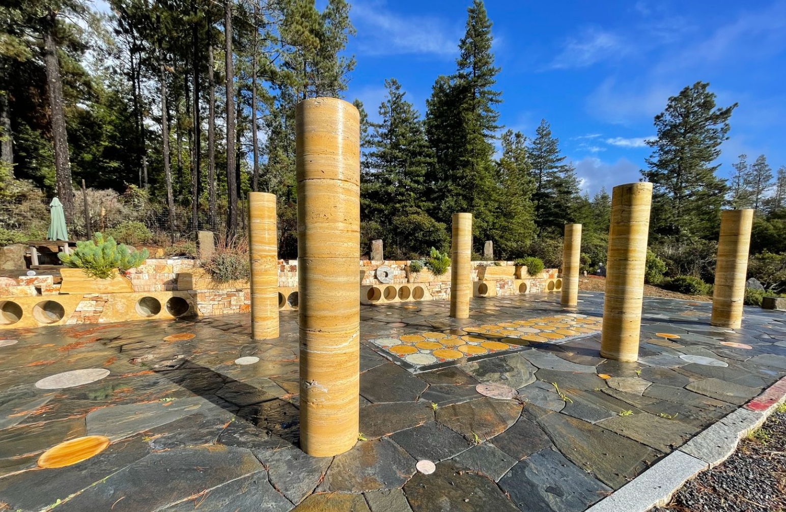 Mendocino Stone Ranch. A stone patio with six tall, cylindrical golden pillars, set against a backdrop of trees and a bright blue sky.