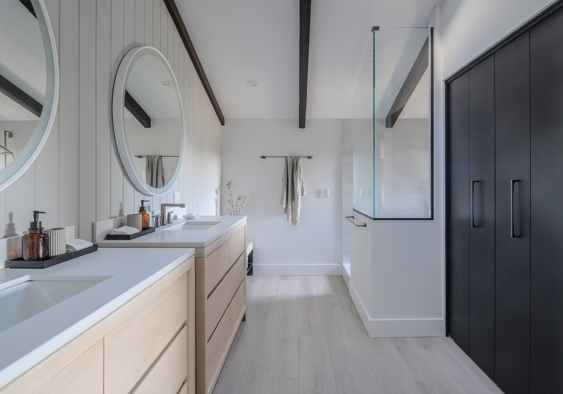 Modern bathroom with double wood vanity, round mirrors, glass walk-in shower, and dark floor-to-ceiling cabinets.