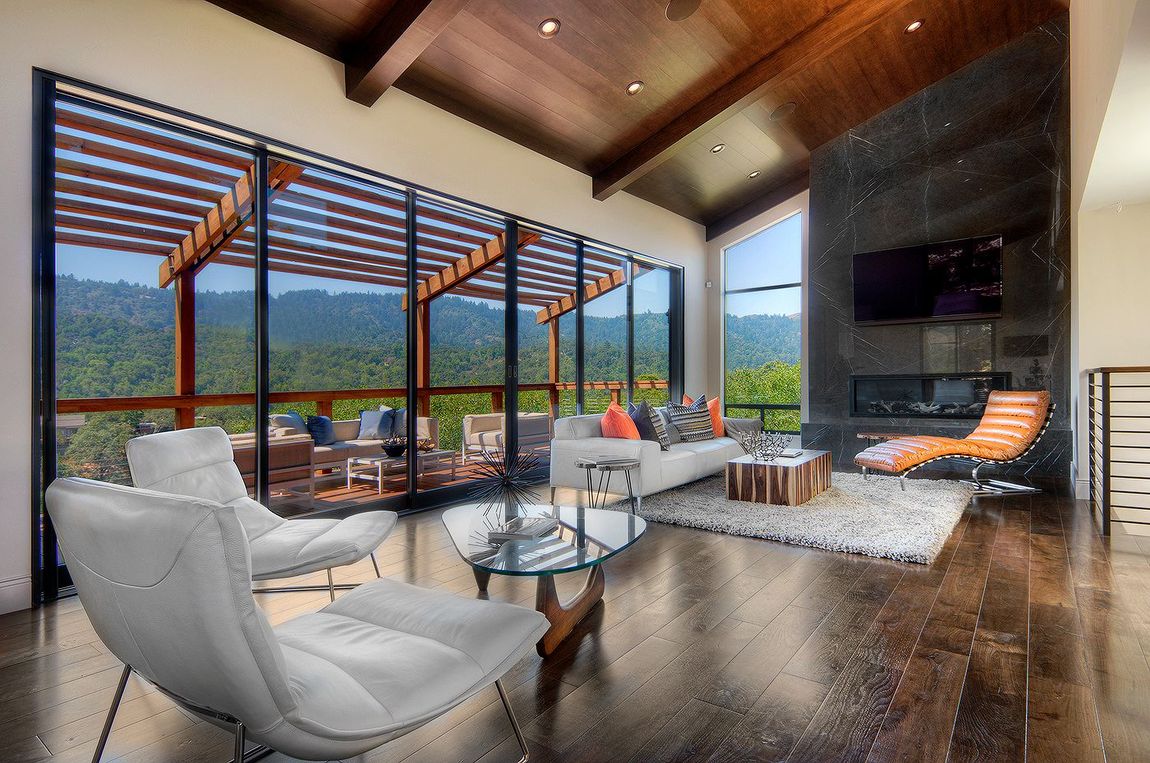 A sea ranch house  living room with hardwood floors, white furniture, a glass coffee table, and a large wall of windows facing trees.