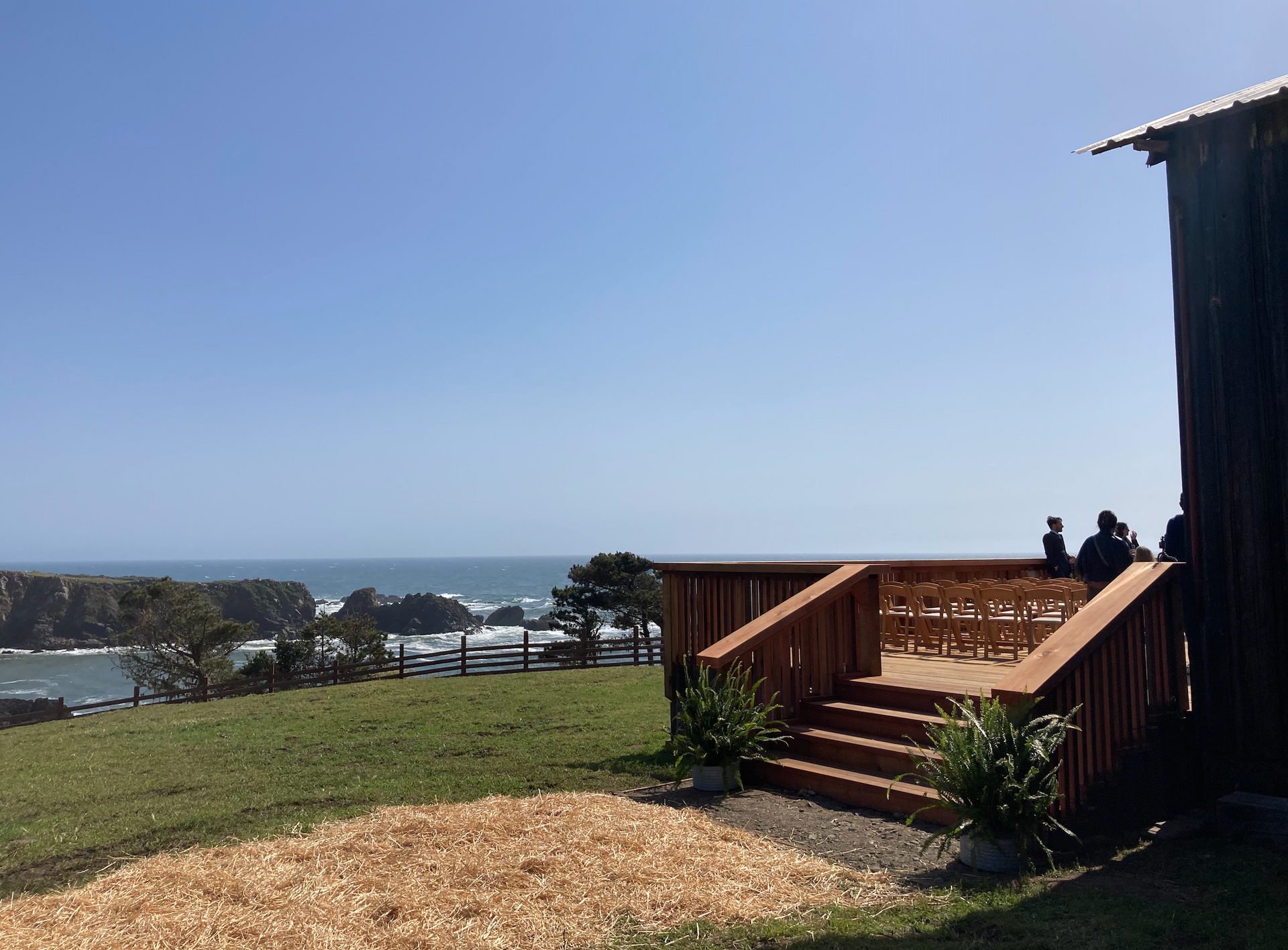The Landing at Stewarts Point. Wedding Barn. A wooden deck overlooks a grassy cliff and the ocean on a bright, sunny day, with a few people standing on the deck.