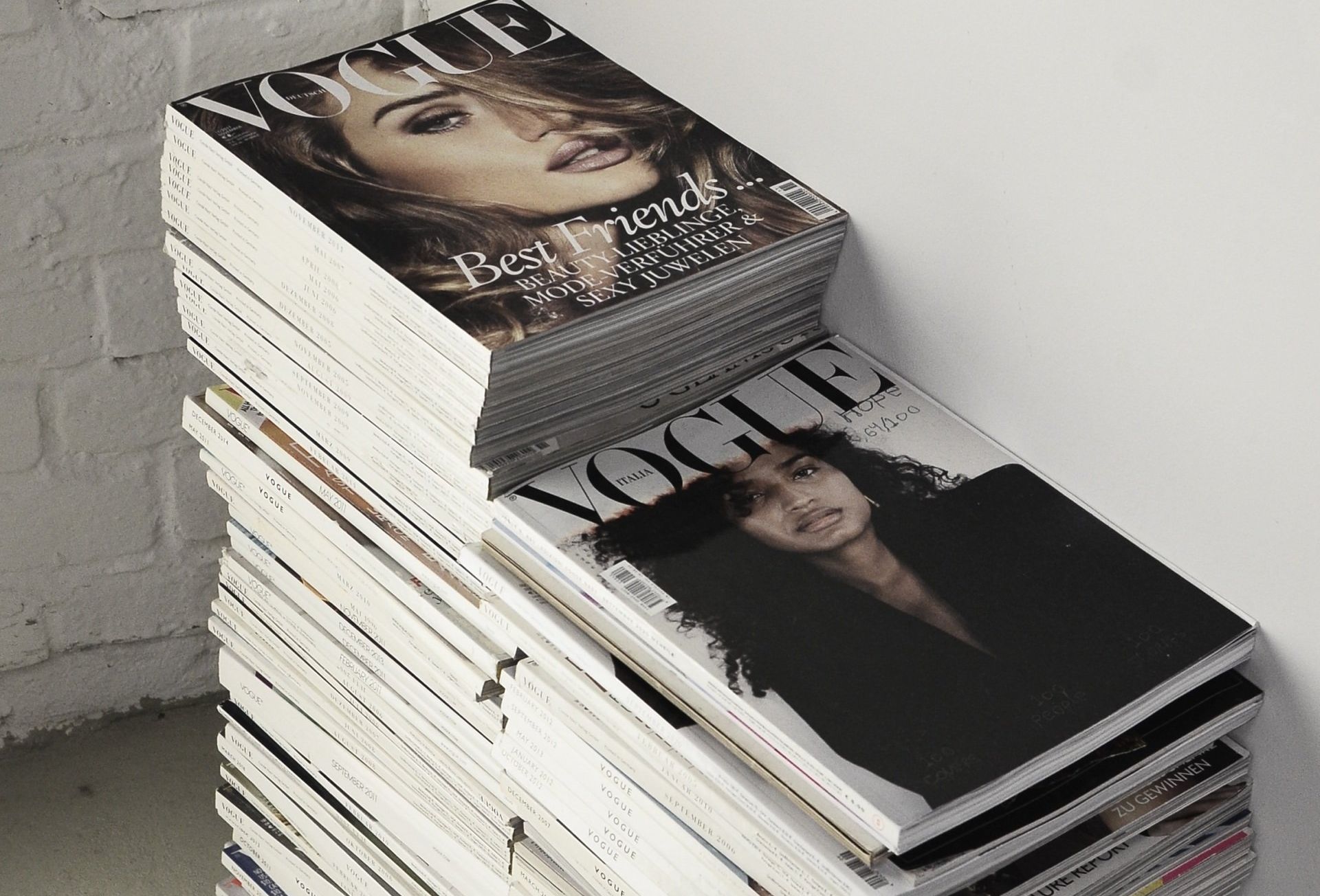 A stack of Vogue magazines sits against a white brick wall.