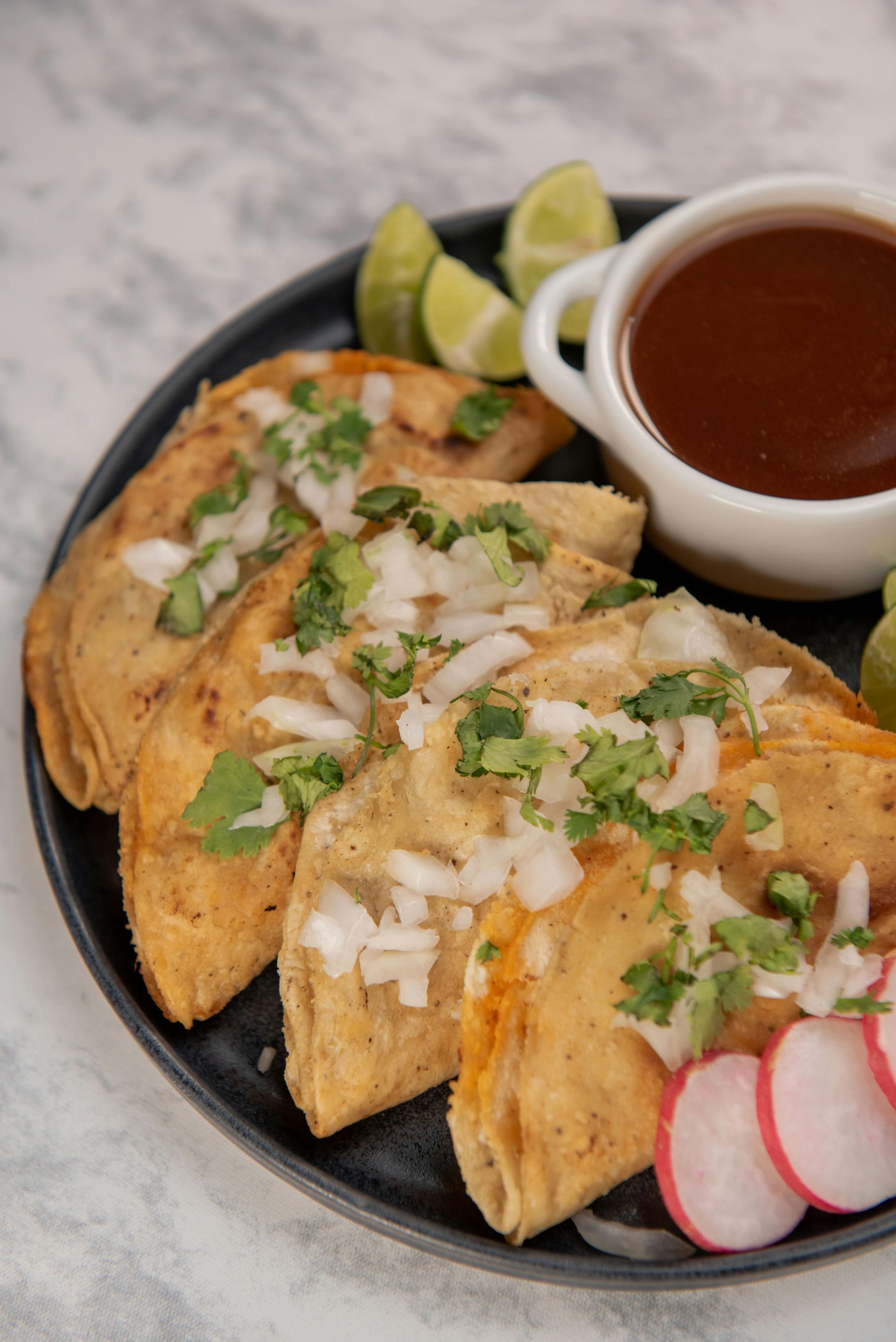 Three birria tacos on a black plate with a side of consommé, lime wedges, and radish slices.