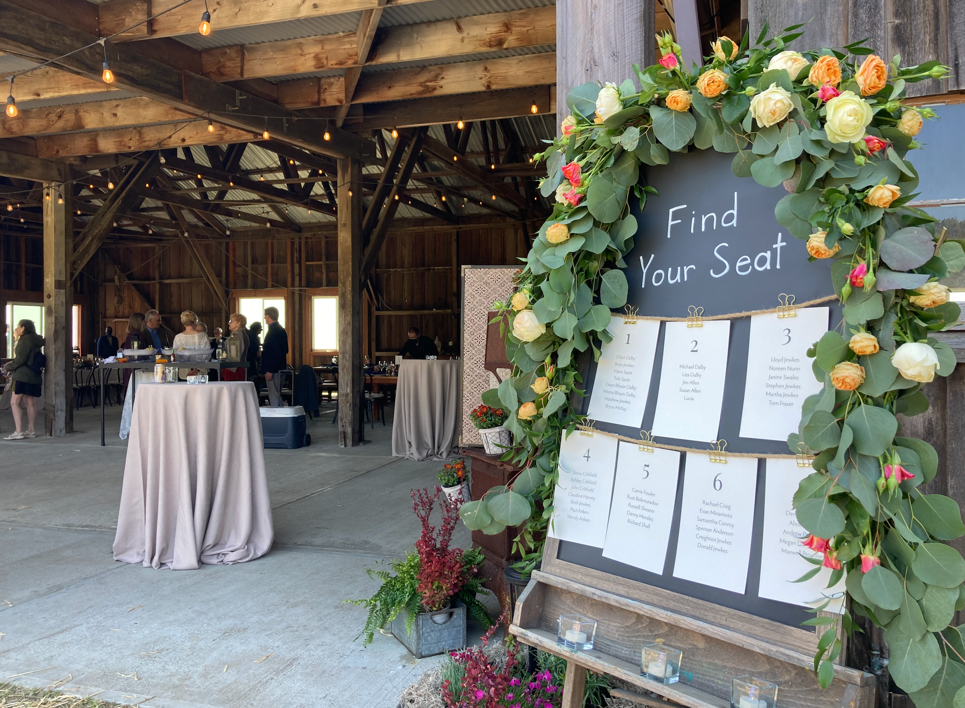 The Landing at Stewarts Point. Wedding Venue. A chalkboard seating chart decorated with flowers and greenery stands inside a rustic barn event space.