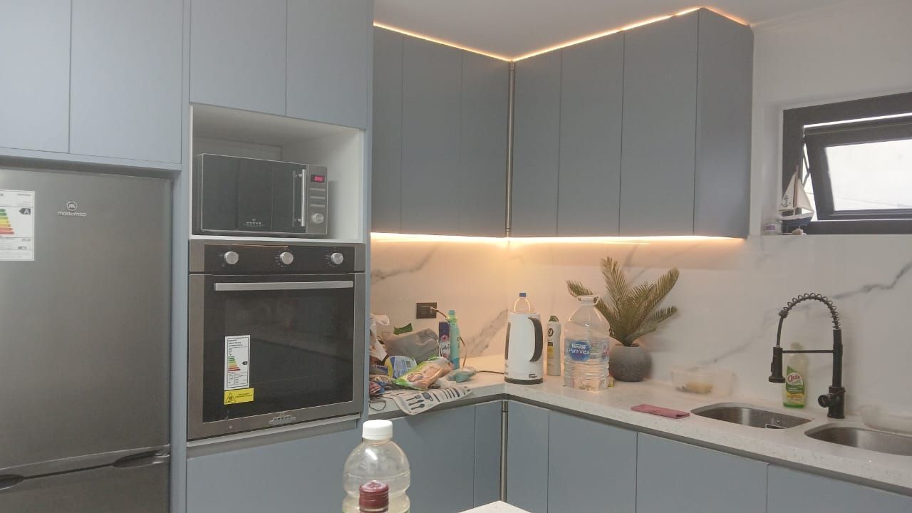 Cocina moderna con gabinetes grises, refrigerador y horno de acero inoxidable, protector contra salpicaduras iluminado y elementos de encimera.