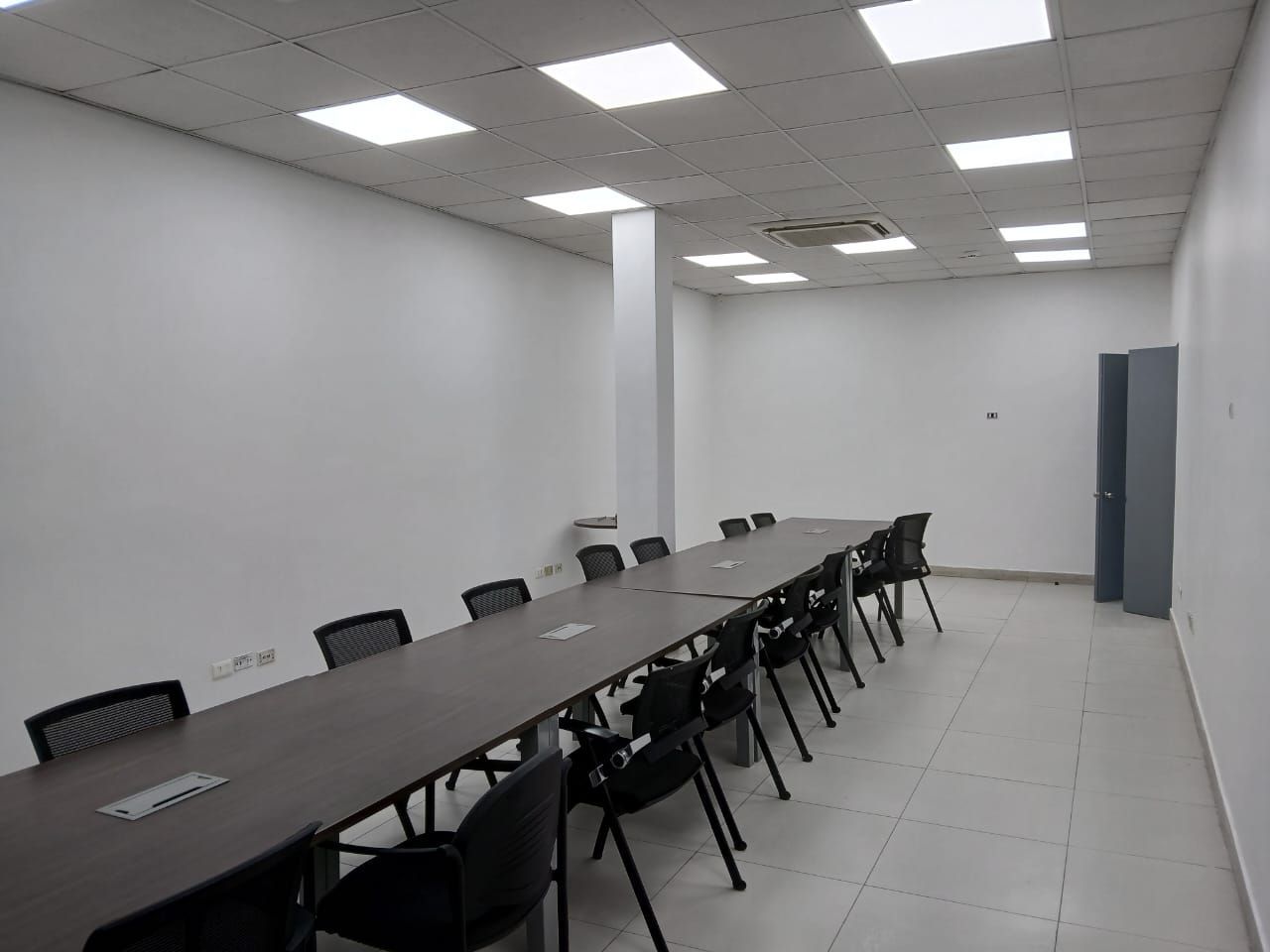 Sala de conferencias vacía con una mesa larga, sillas negras y paredes blancas bajo luces fluorescentes en el techo.