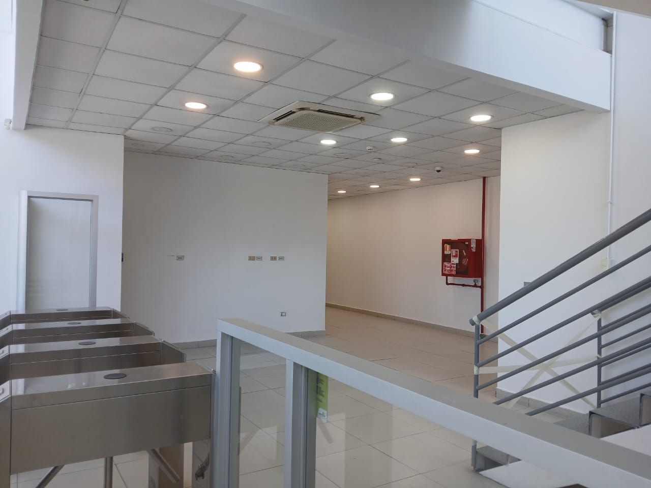 Interior blanco vacío con luces de techo de azulejos, una barandilla de escalera y una puerta abierta a la izquierda.