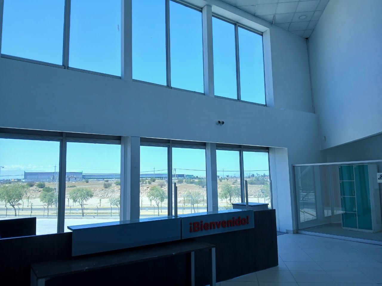 Terminal de aeropuerto vacía con grandes ventanales y un mostrador de facturación bajo una brillante luz natural.
