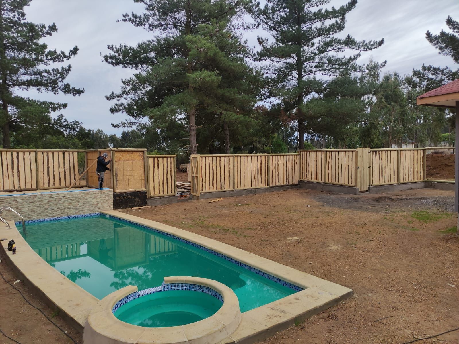 Piscina y jacuzzi en el patio trasero, junto a una valla de madera y pinos.