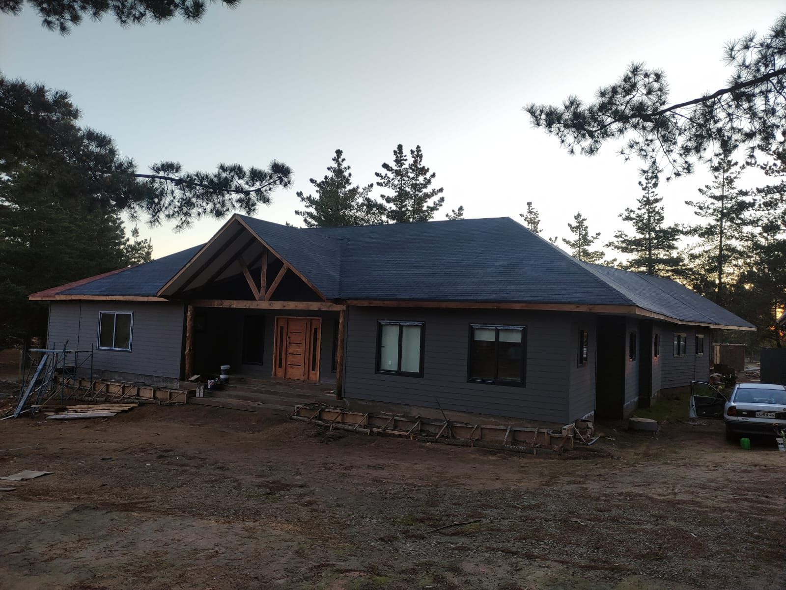 Casa gris en construcción al anochecer, con techo oscuro, marco de entrada frontal y árboles a su alrededor.
