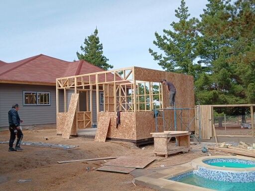 Cobertizo de madera parcialmente construido junto a una casa, con piscina y un trabajador en una obra en construcción en el patio trasero.