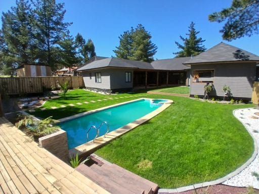 Patio trasero con piscina turquesa, césped verde y casa gris bajo un cielo azul claro.
