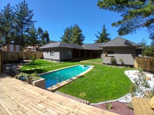 Patio trasero con piscina, césped verde, terraza de madera y casa de una sola planta bajo un cielo azul.
