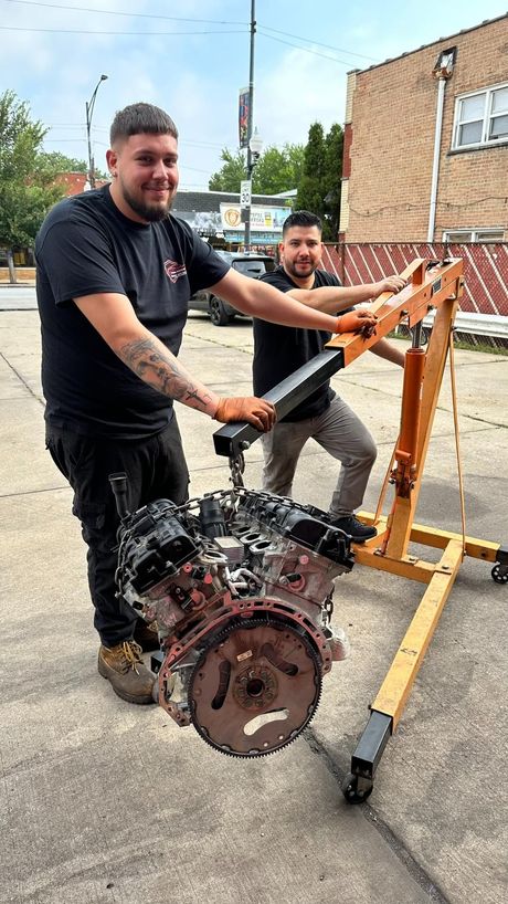 Two men posing with a large engine mounted on a wooden stand outdoors.