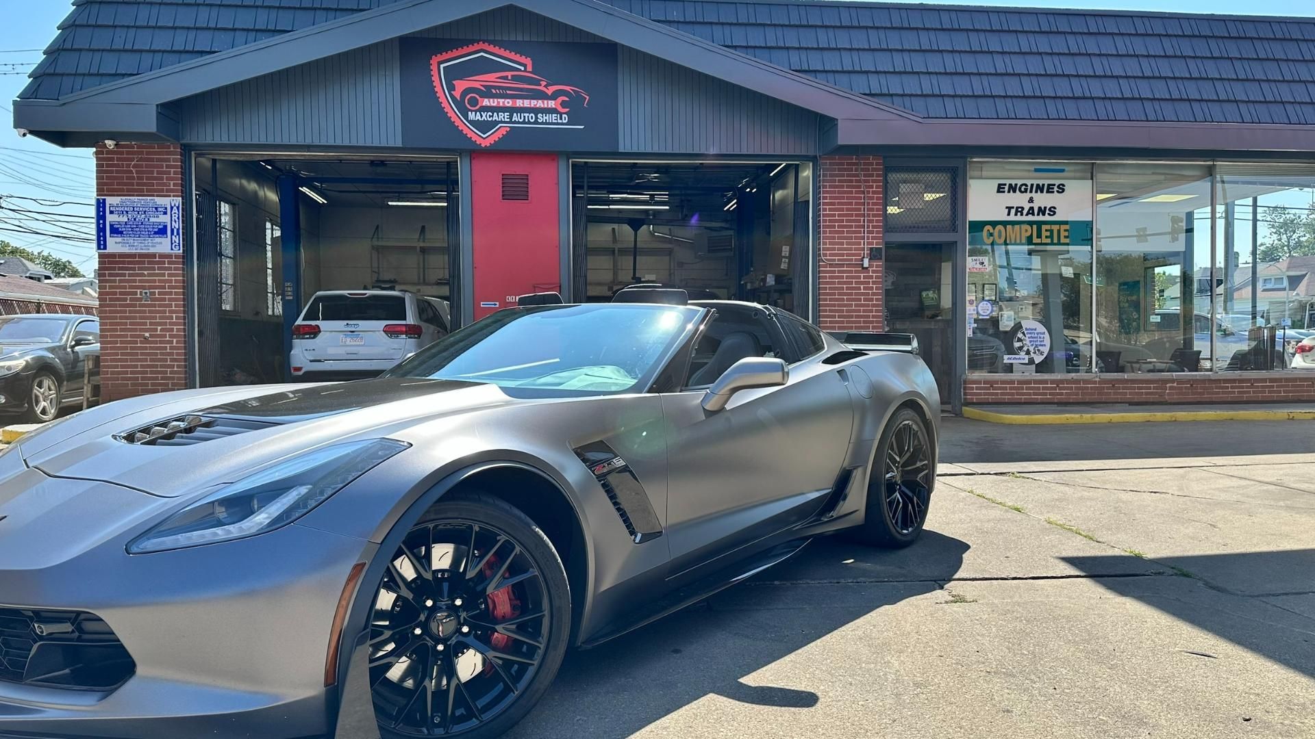 Gray sports car parked outside a car dealership with a neon sign and storefront windows